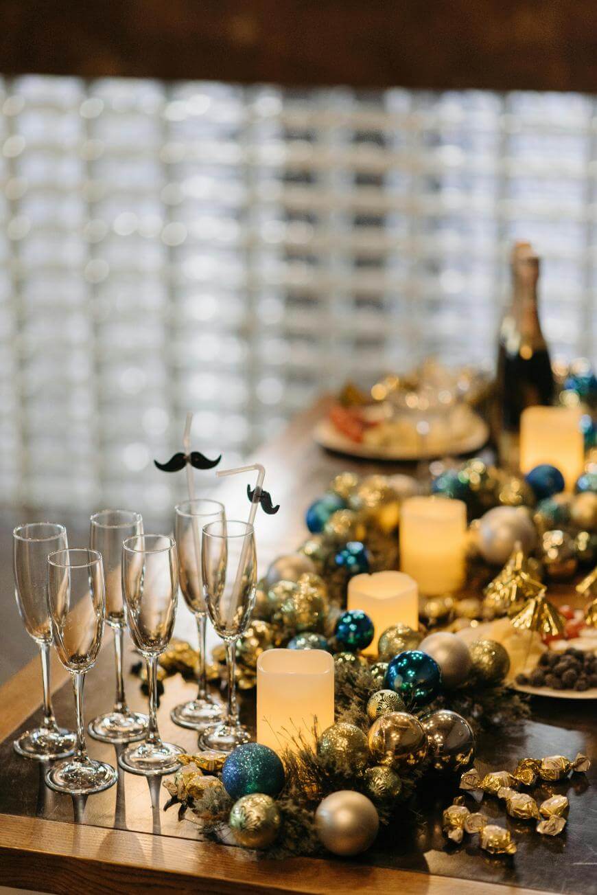 A festive table setting with champagne flutes, decorative candles, gold and blue ornaments, a champagne bottle, food platters, and two glasses topped with mustache-shaped drink markers.