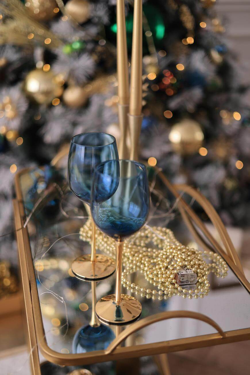 Two blue wine glasses with gold stems sit on a gold tray with a strand of pearls, in front of a decorated Christmas tree with lights, gold ornaments, and blurred festive background.
