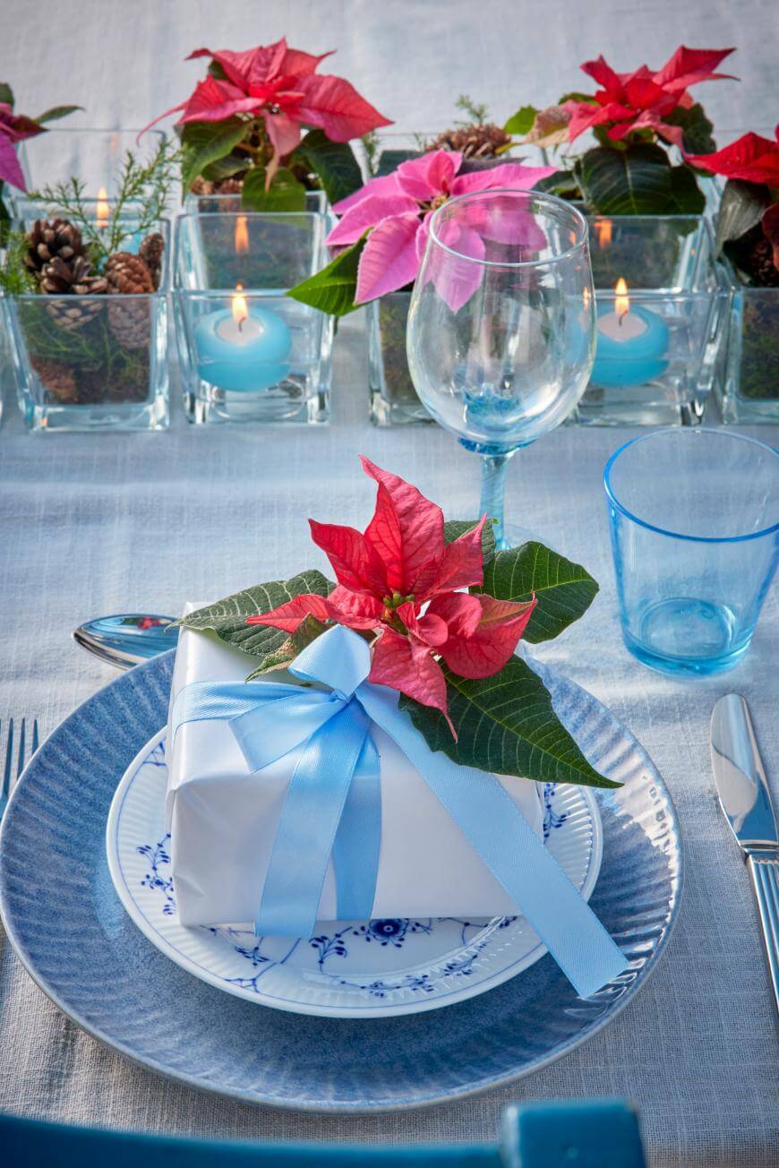 A festive table setting with a wrapped gift topped with a red poinsettia and blue ribbon on a plate, surrounded by glassware, silverware, candles, pine cones, and poinsettia centerpieces.