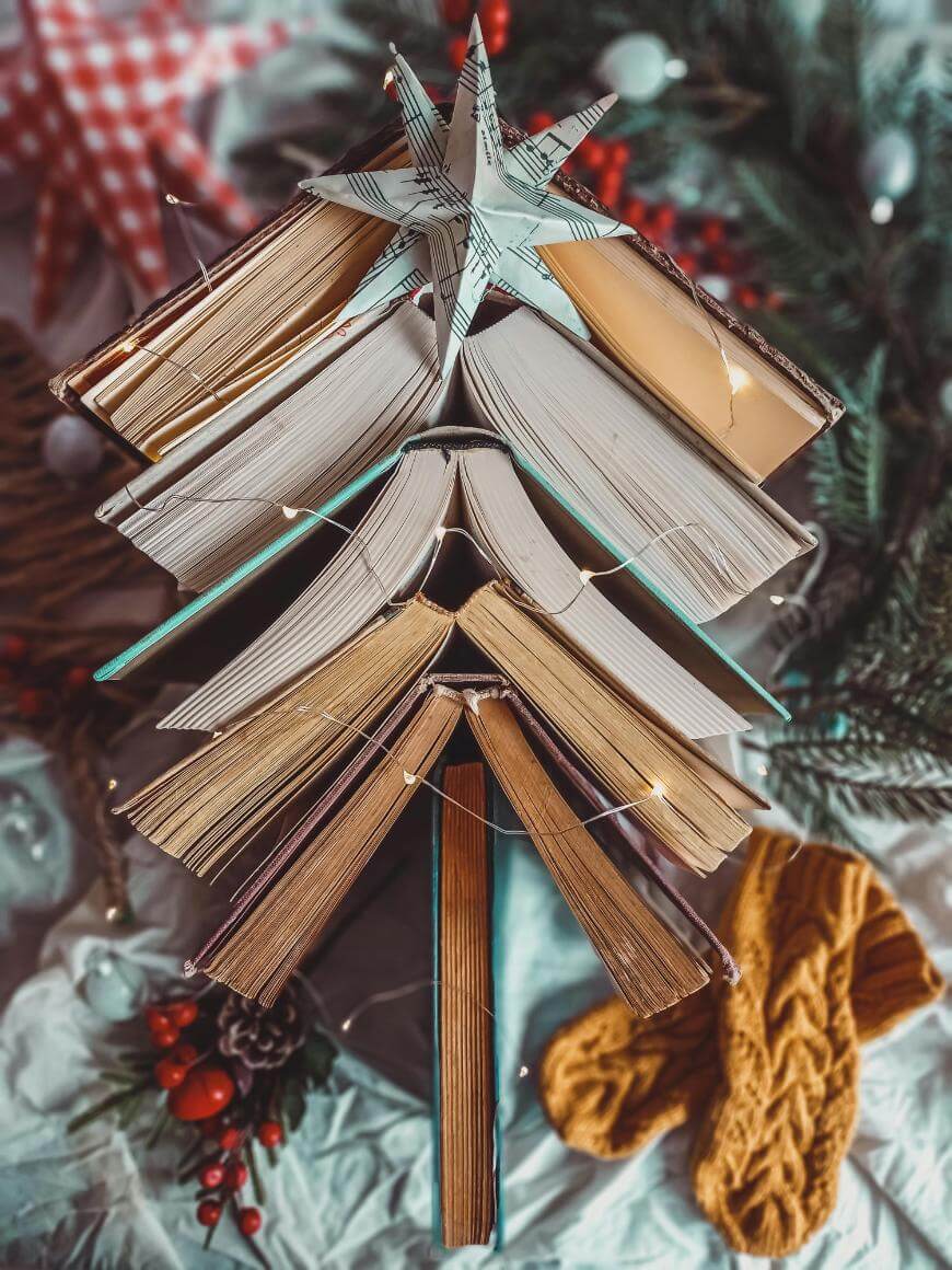 A stack of open books arranged in the shape of a Christmas tree, topped with a star and decorated with string lights, surrounded by festive decorations and a pair of yellow knitted mittens.