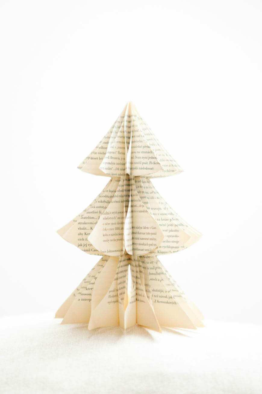 A small, stylized Christmas tree made from folded book pages stands upright against a plain white background. The tree has layered, triangular sections resembling branches.