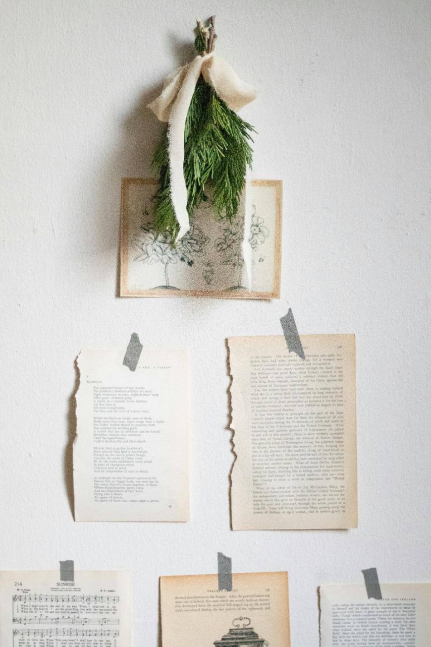 A small bundle of evergreen branches tied with white fabric hangs on a wall above three vintage book pages taped to the wall with washi tape, creating a simple, artistic display.