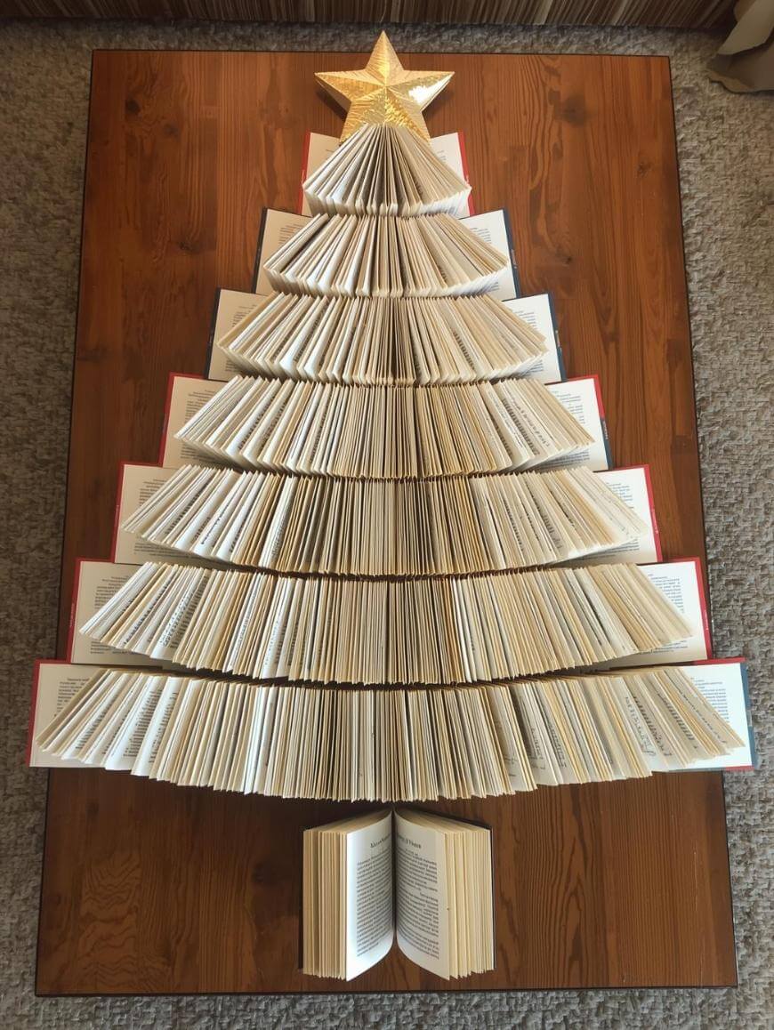 Books arranged on a wooden table in the shape of a Christmas tree, with open pages fanned out and a golden star decoration at the top. An open book sits at the bottom as the tree trunk.