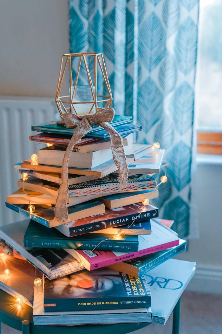 A stack of books arranged in a tree shape, decorated with a burlap ribbon, golden geometric ornament on top, and string lights, in front of a window with blue patterned curtains.