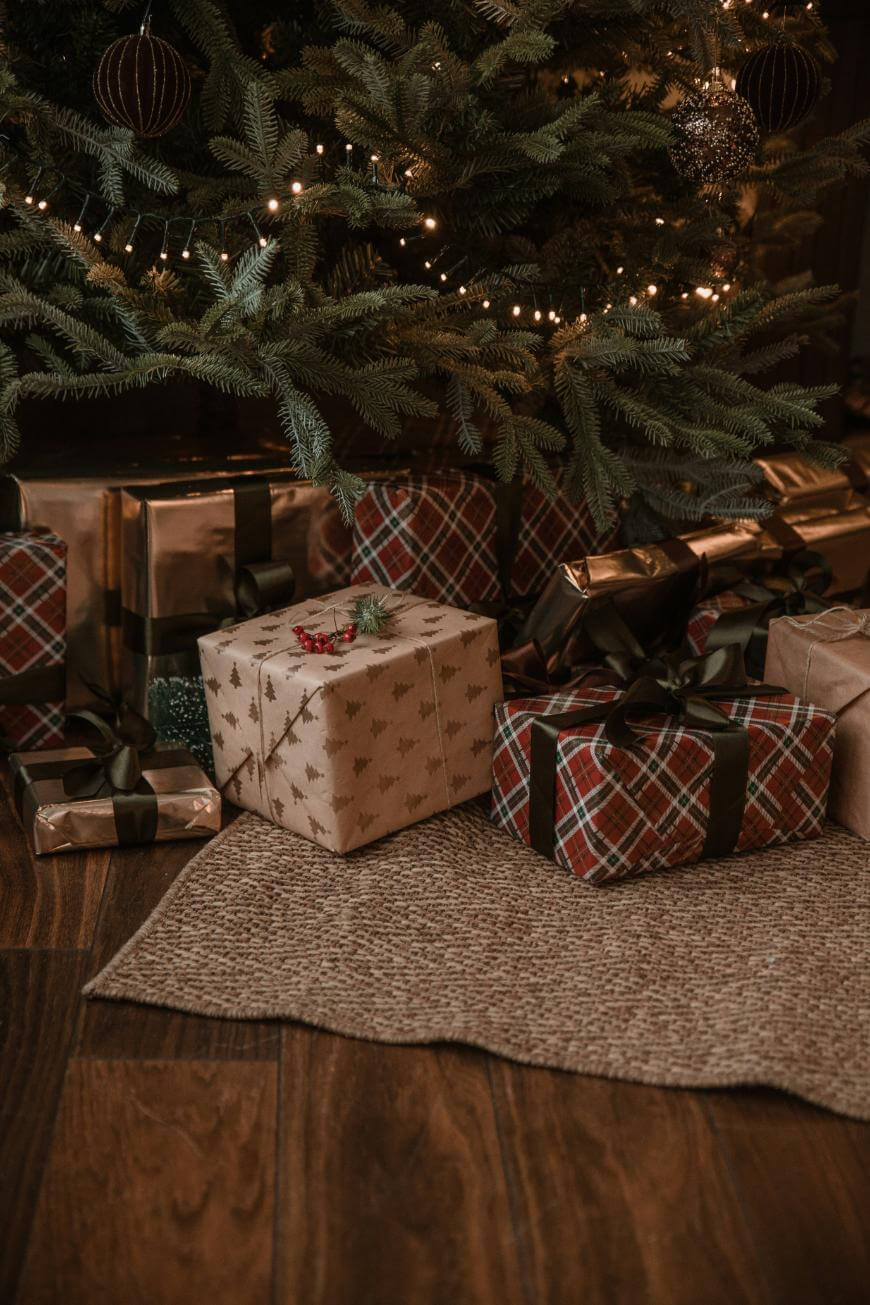 Several wrapped Christmas presents with ribbons are placed under a decorated Christmas tree with glowing lights, sitting on a wooden floor next to a textured rug.