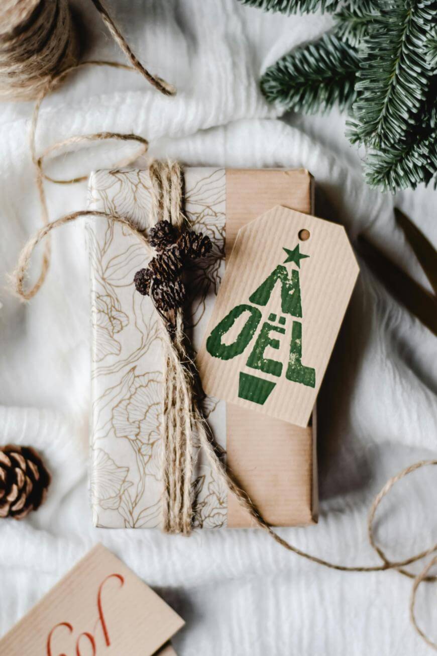 A gift wrapped in patterned paper is tied with twine and decorated with a pinecone. A tag with a green Christmas tree and NOËL hangs from the twine. Pine branches and ribbon are nearby.
