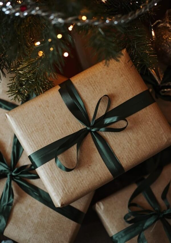 A close-up of gifts wrapped in brown paper and tied with dark green ribbons, placed under a decorated Christmas tree with lights.