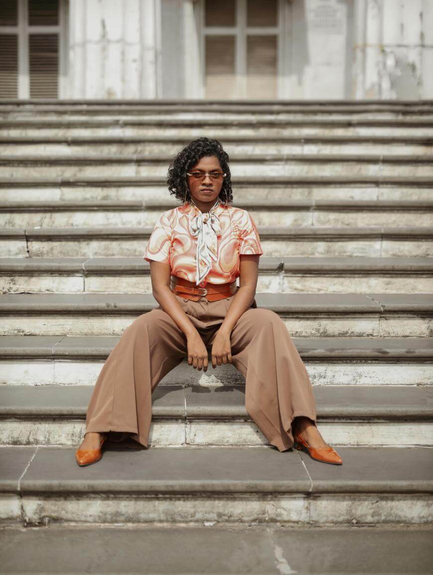 Person with curly hair wearing glasses, a patterned blouse, brown wide-leg pants, and orange shoes sits confidently on outdoor stone steps, looking directly at the camera.