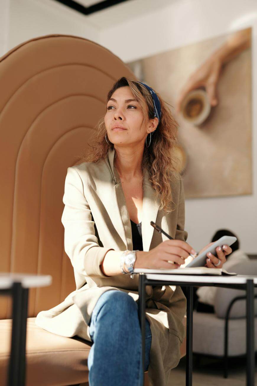 A woman with wavy hair, wearing a beige blazer and blue jeans, sits on a tan chair, holding a pen and notepad, looking thoughtfully to the side in a bright, modern room.