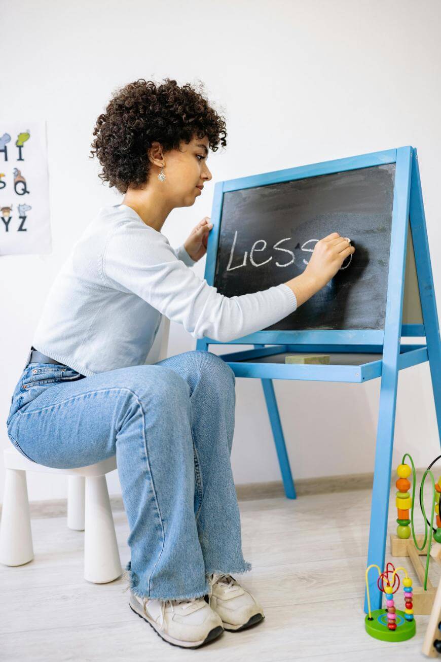 A young woman with curly hair writes the word “Lesen” on a blackboard with chalk. She is seated on a stool in a bright room, with educational toys and an alphabet chart visible nearby.