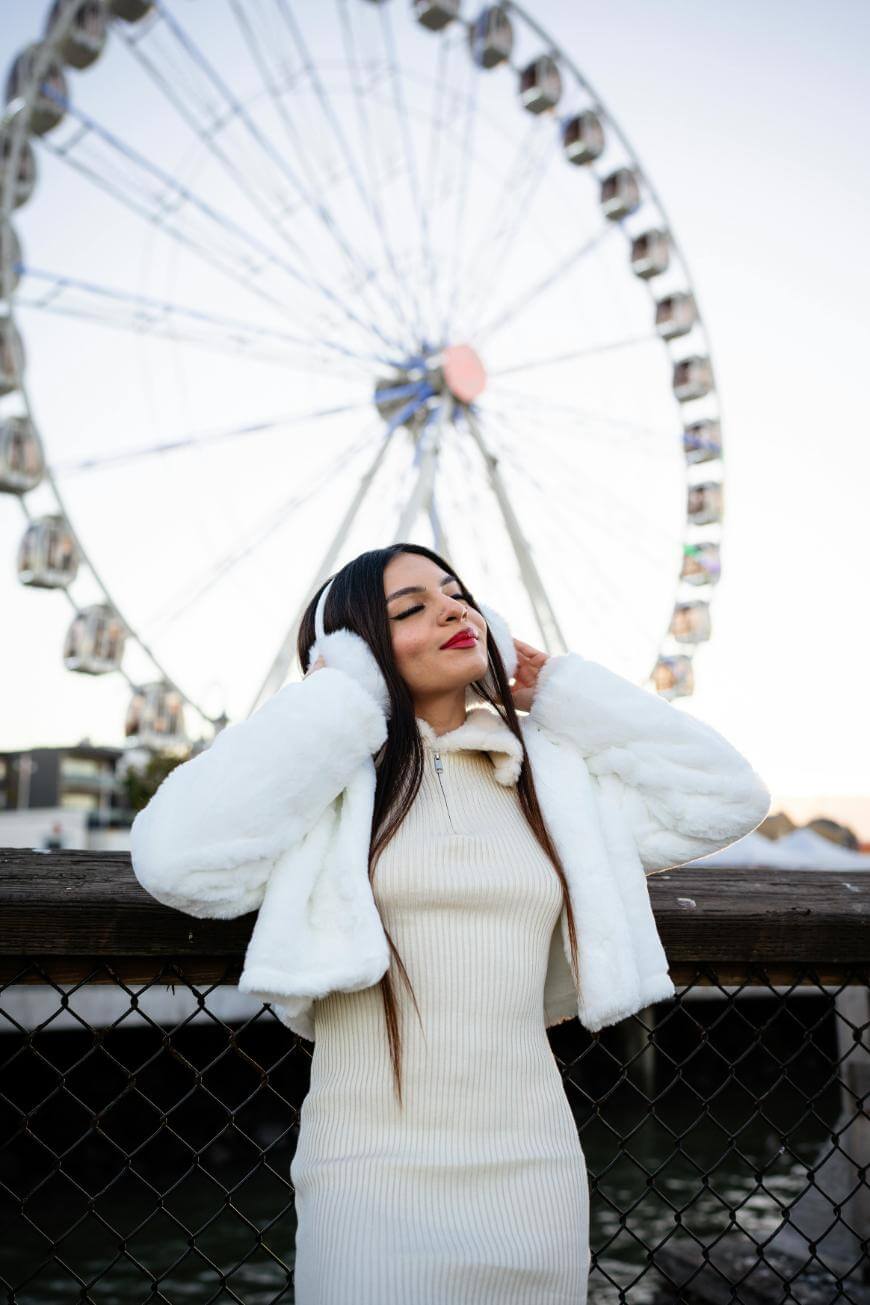 A woman in a white coat and dress stands by a fence, listening to headphones with her eyes closed. Behind her is a large Ferris wheel against a clear sky.