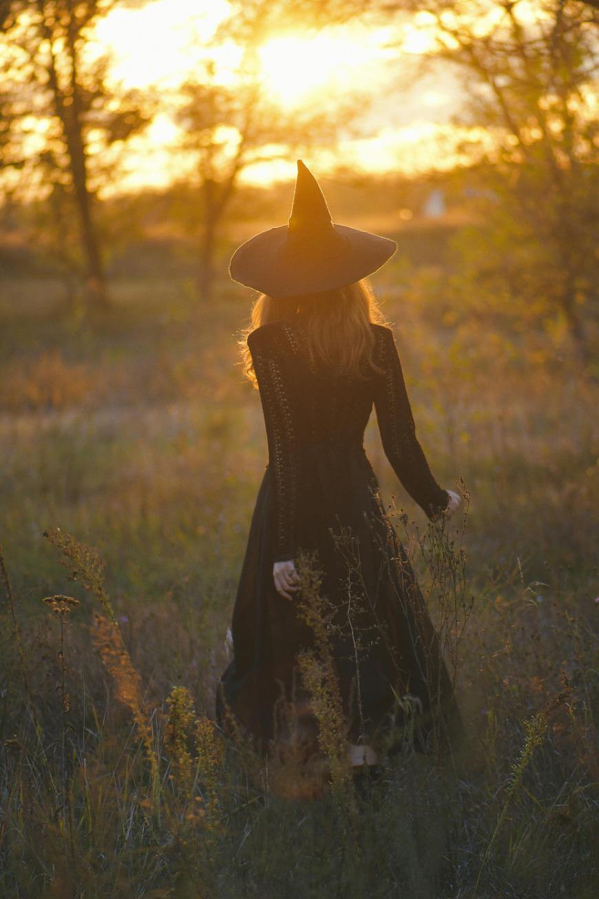 A person in a black dress and pointed witch hat walks through a grassy field at sunset, with golden sunlight streaming through the trees in the background.