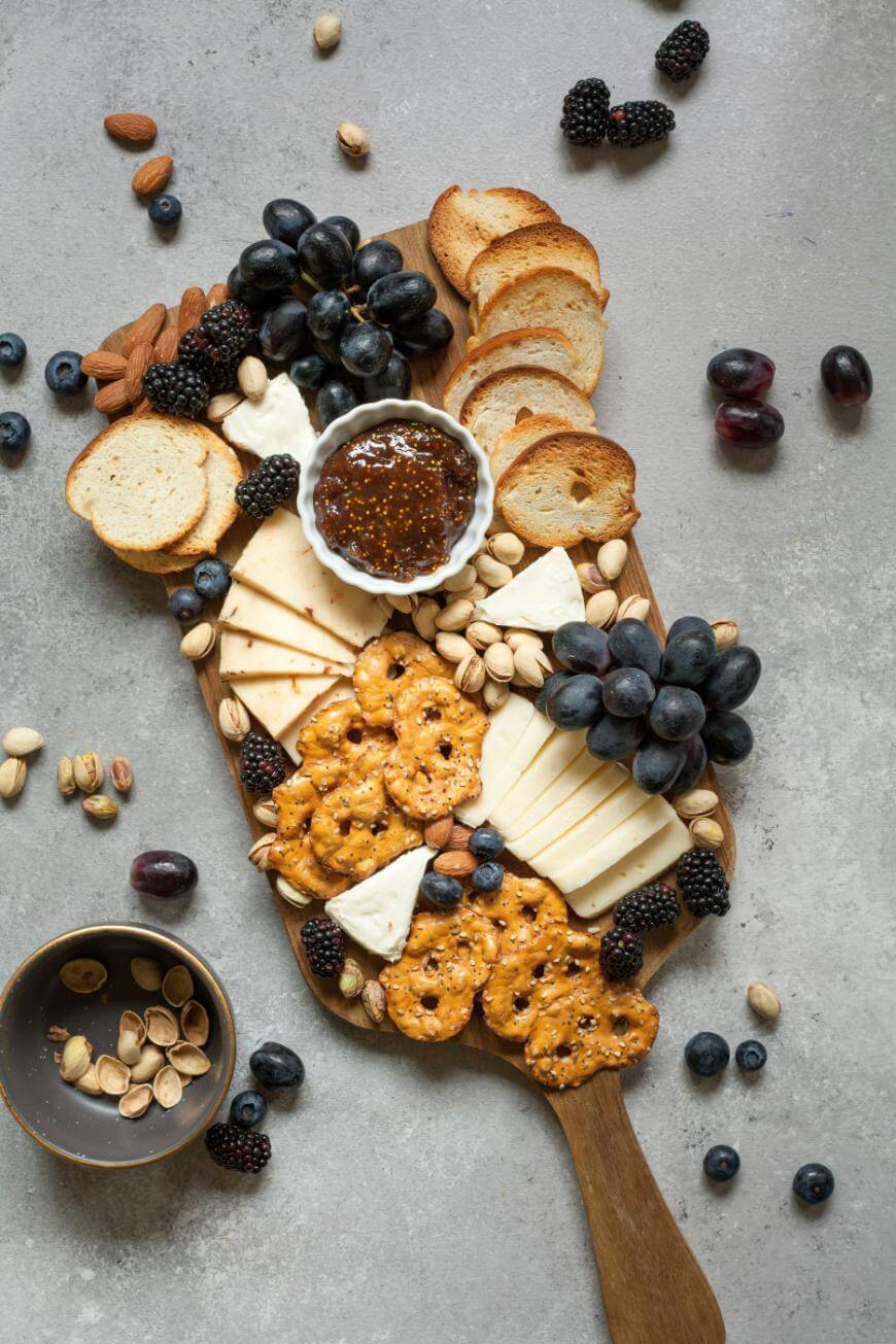A wooden serving board with assorted cheeses, pretzels, sliced baguette, blackberries, grapes, nuts, and a small bowl of jam, with scattered fruit and nuts on a gray surface.