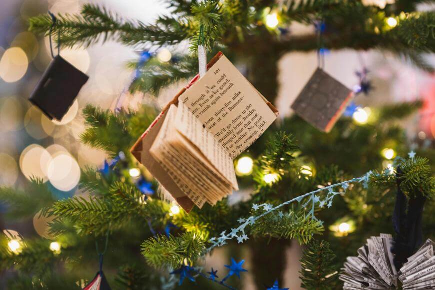 A Christmas tree decorated with string lights, blue star garland, and handmade ornaments resembling tiny open books made from printed pages.