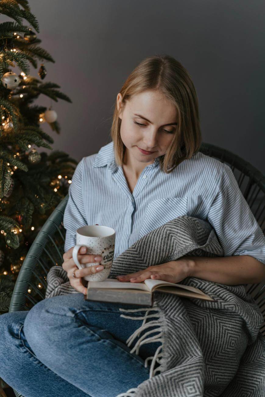 A young woman sits in a chair by a decorated Christmas tree, wrapped in a gray blanket, holding a mug, and reading a book. She has short blonde hair and wears a striped shirt and jeans.