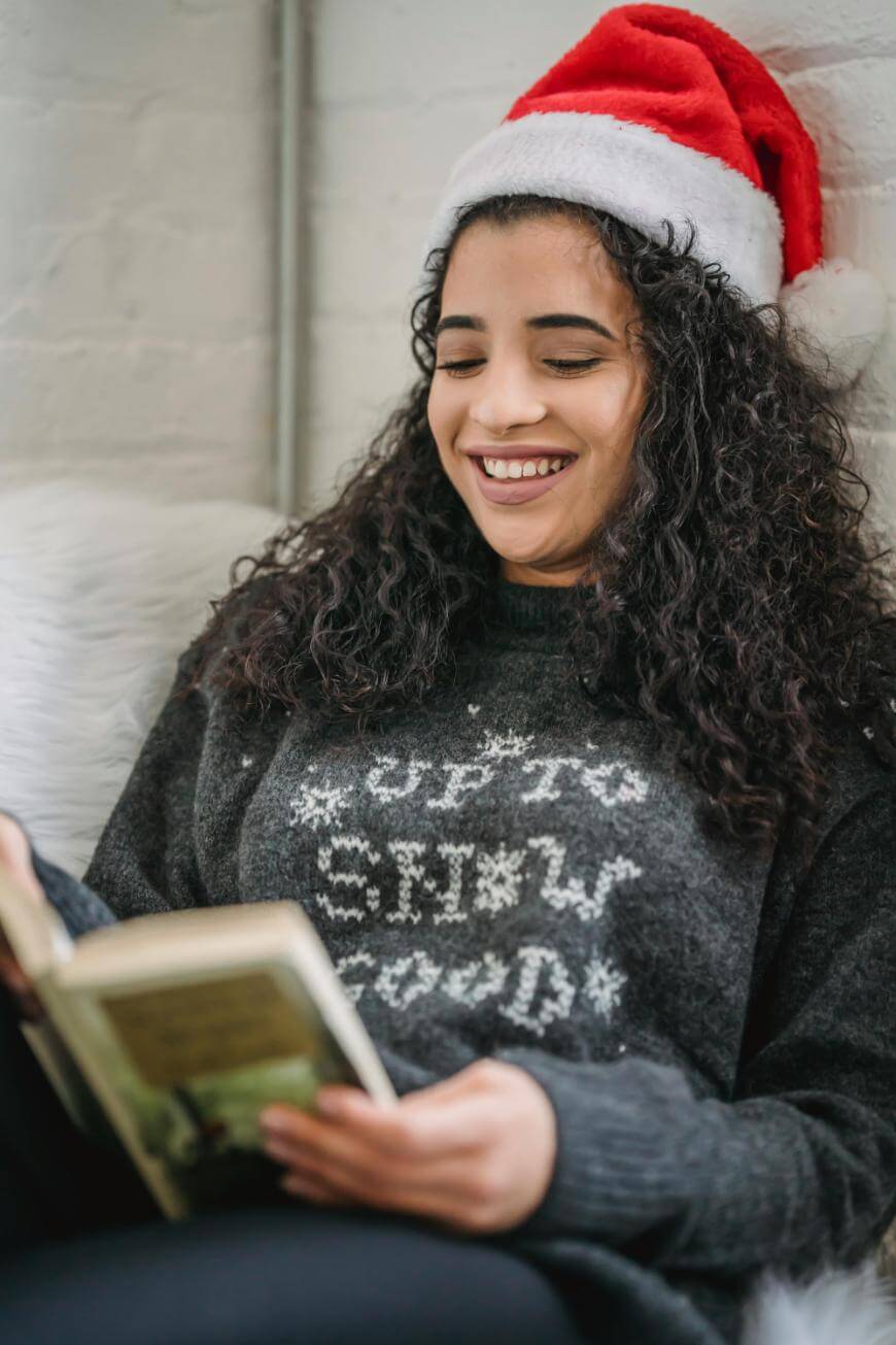 A smiling woman with curly hair, wearing a Santa hat and a festive sweater, sits and reads a book.
