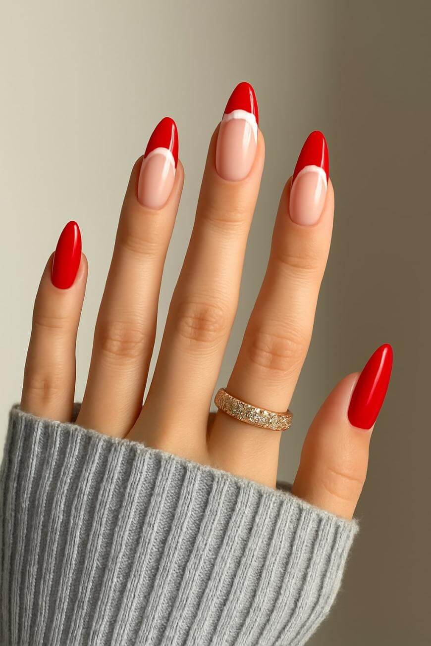 A hand with long, almond-shaped nails painted in a red and nude French tip design, wearing a gold ring with small diamonds, and partially covered by a gray ribbed sweater.