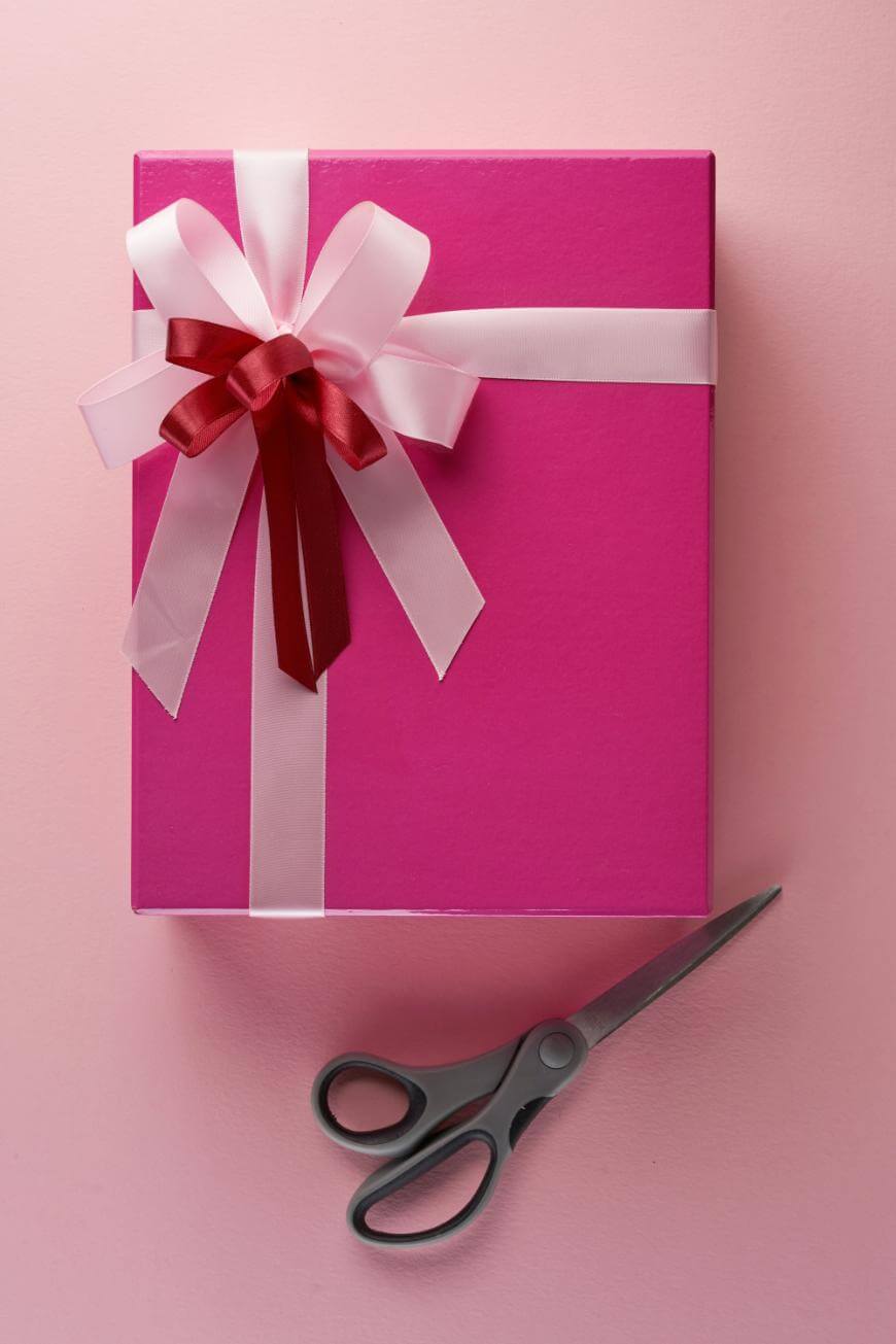 A pink gift box with white and red ribbon bows sits on a pink surface. A pair of black-handled scissors is placed below the box.