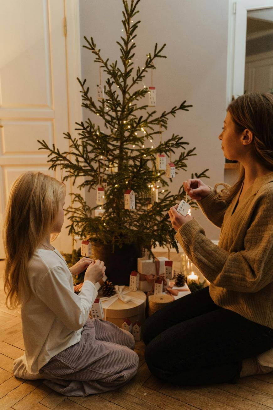 A woman and a young girl kneel by a decorated Christmas tree with string lights and tags, placing ornaments. Wrapped presents and pinecones are arranged at the base of the tree in a cozy indoor setting.