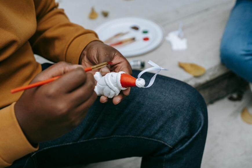 A person is painting a small white object with red paint using a brush. Paint supplies and dried leaves are visible in the background on a light surface.