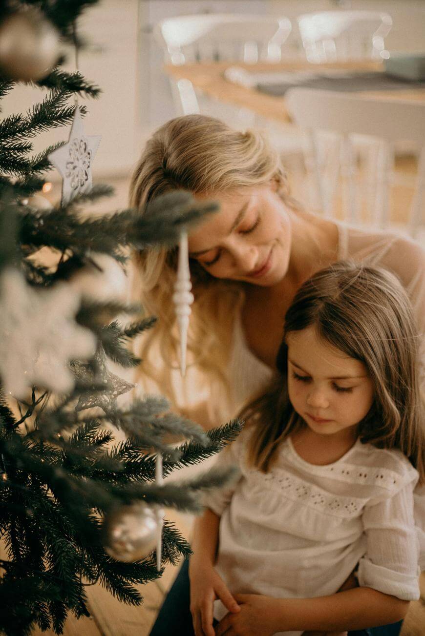 A woman and a young girl sit closely together beside a decorated Christmas tree, both gazing down with peaceful expressions. The tree is adorned with ornaments and the setting feels warm and festive.