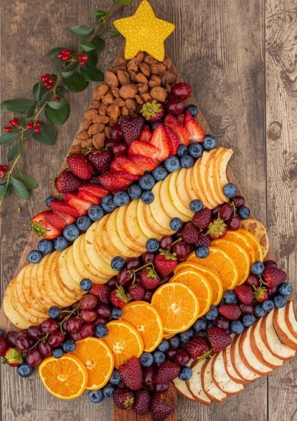 A wooden board arranged with fruit, cheese, and nuts in the shape of a Christmas tree, topped with a yellow star and decorated with berries, sitting on a rustic wooden surface.