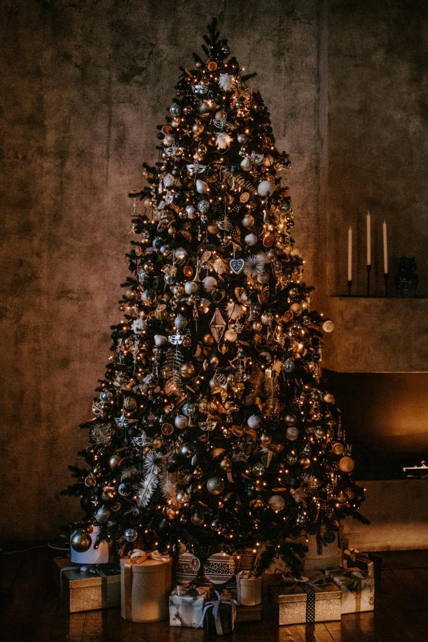 A tall, beautifully decorated Christmas tree adorned with gold and silver ornaments and warm string lights stands in a dimly lit room, with wrapped presents arranged at the base and candles on a shelf nearby.