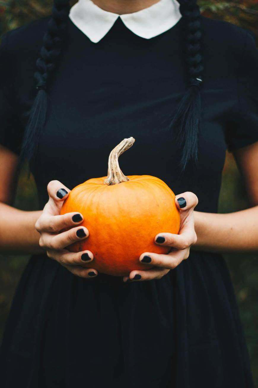A person wearing a black dress with a white collar, and black nail polish, holds a small orange pumpkin with both hands. The persons braided hair and upper body are visible, but their face is not shown.