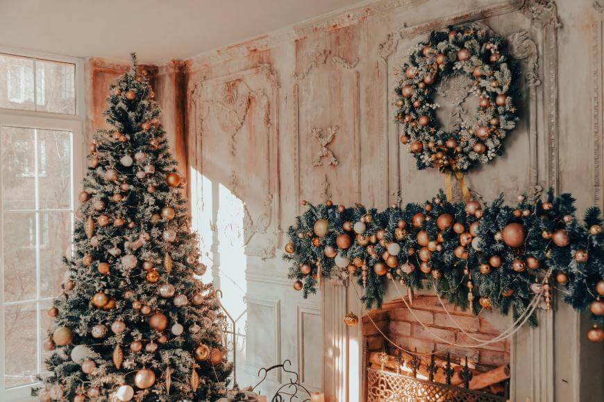 A decorated Christmas tree stands beside a fireplace adorned with garlands and a large wreath. Gold and white ornaments and warm sunlight create a festive, cozy atmosphere in the elegant room.