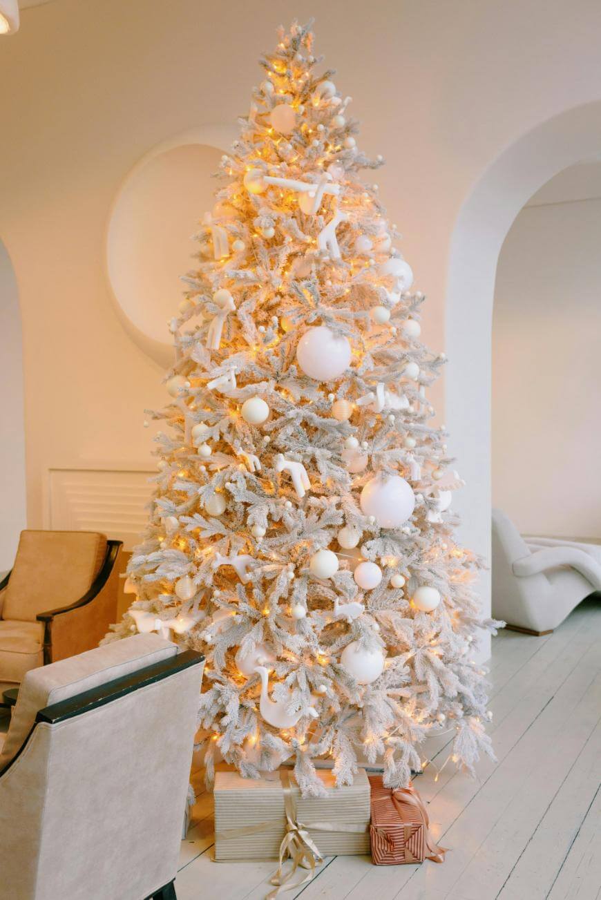 A large, white Christmas tree decorated with white ornaments and warm lights stands in a bright, modern living room with neutral furniture and wrapped presents underneath.