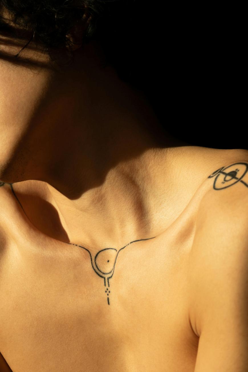 A close-up of a persons bare upper chest and shoulder, featuring minimalist black line tattoos, including one resembling a stylized uterus on the chest and another abstract design on the upper arm, against a dark background.