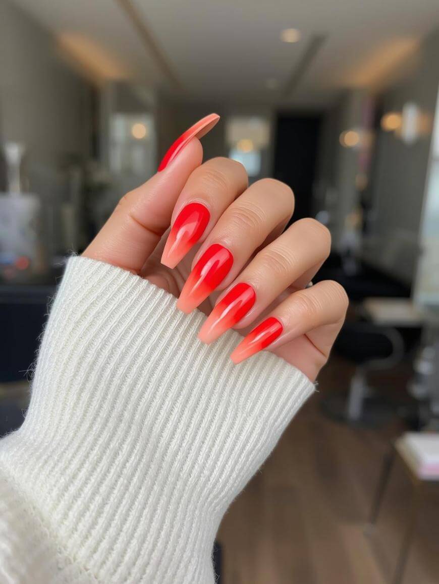 A hand with long, almond-shaped nails featuring a red and peach ombre design is shown. The person is wearing a white knit sweater, and the background appears to be a brightly lit modern salon.