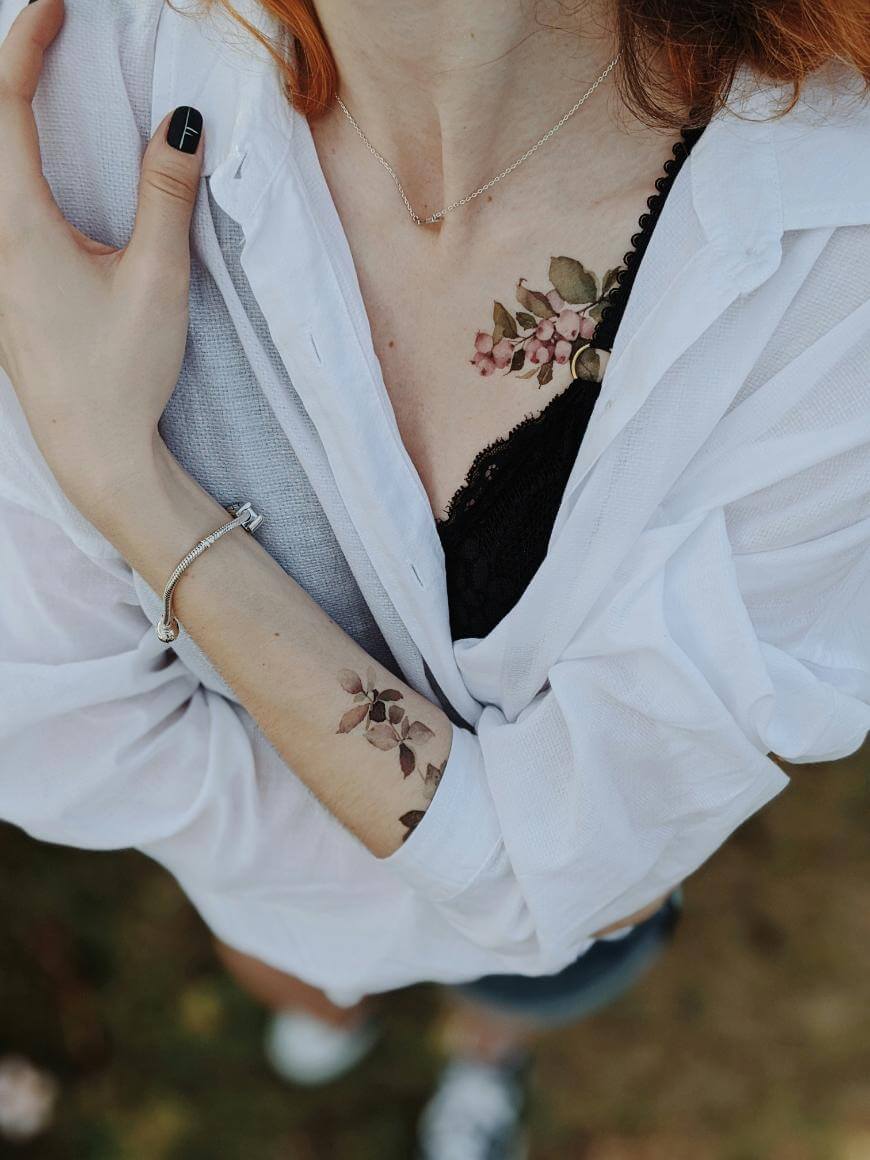 A woman wearing a white shirt and black lace top shows floral tattoos on her chest and forearm. She has a silver bracelet and necklace, and her nails are painted black. The ground with grass is visible below her.