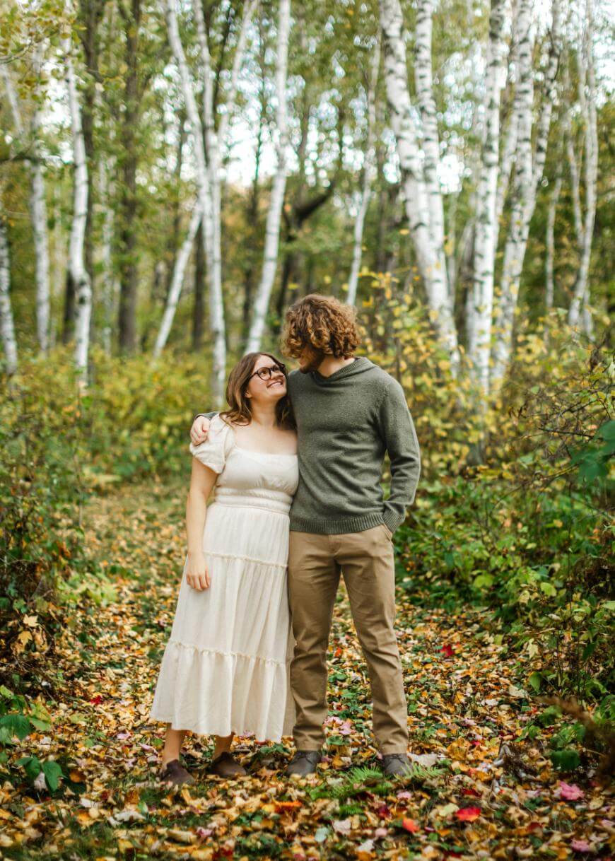 A couple stands close together on a leafy path in a birch forest, smiling at each other. The woman wears a white dress, and the man wears a green sweater and tan pants. It is a bright, peaceful autumn day.