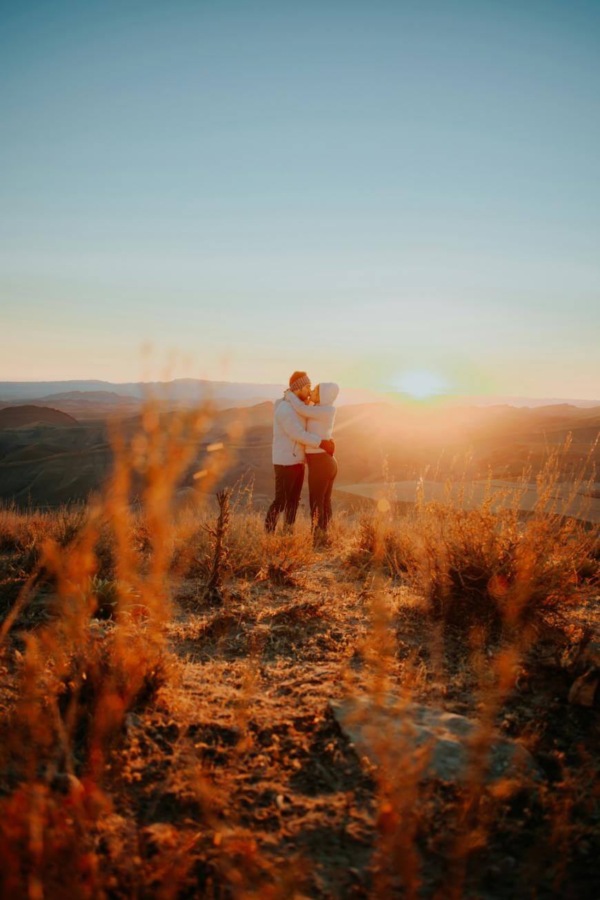 A couple embraces on a grassy hilltop at sunset, surrounded by golden light and dry vegetation, with distant mountains and a clear sky in the background.