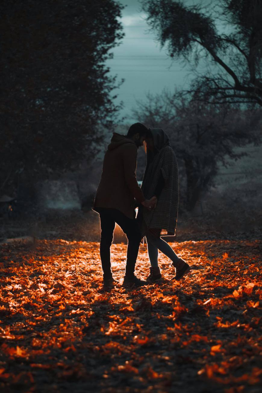 A couple stands close together, holding hands and sharing an intimate moment, surrounded by fallen autumn leaves on a dimly lit path, with dark trees and a moody sky in the background.