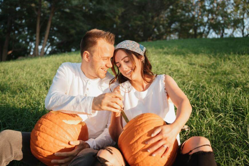 A smiling couple sits on grass, carving pumpkins together in sunlight. The woman holds a knife and pumpkin, while the man watches her, both enjoying a relaxed outdoor moment. Trees and greenery are in the background.