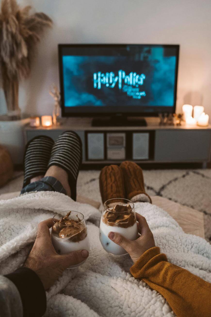 Two people sitting on a couch under a blanket, holding dessert glasses, with a TV in front of them displaying the Harry Potter movie logo. The room is cozy with candles and soft lighting.
