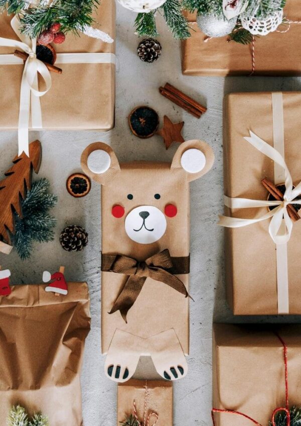 A collection of gifts wrapped in brown paper and tied with ribbons, with one present decorated as a cute bear with paper ears, paws, rosy cheeks, and a bow, surrounded by festive holiday ornaments and greenery.