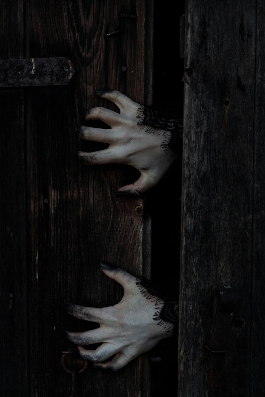 Two pale hands with black-painted fingertips and lace cuffs reach through the narrow opening of a dark, weathered wooden door, creating an eerie and mysterious atmosphere.