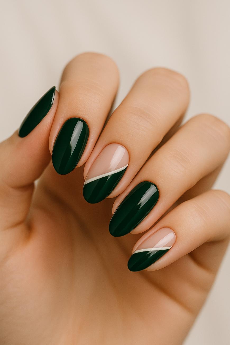 A hand with almond-shaped nails features a modern manicure; most nails are painted dark green, while two have a diagonal nude and green design. The background is a soft, light color.