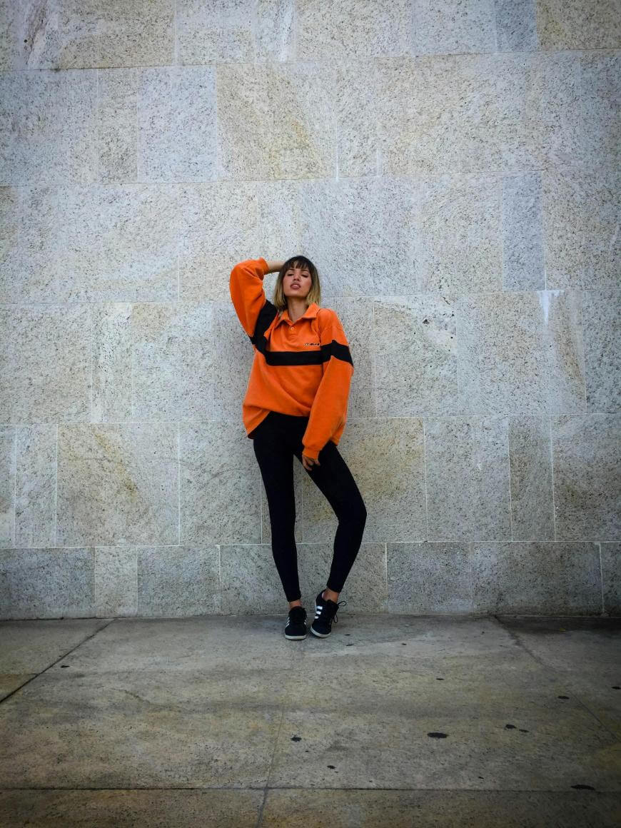 A person stands against a pale stone wall, wearing a loose orange sweater with a black stripe, black pants, and black shoes. One hand is touching their head, and they are looking at the camera.