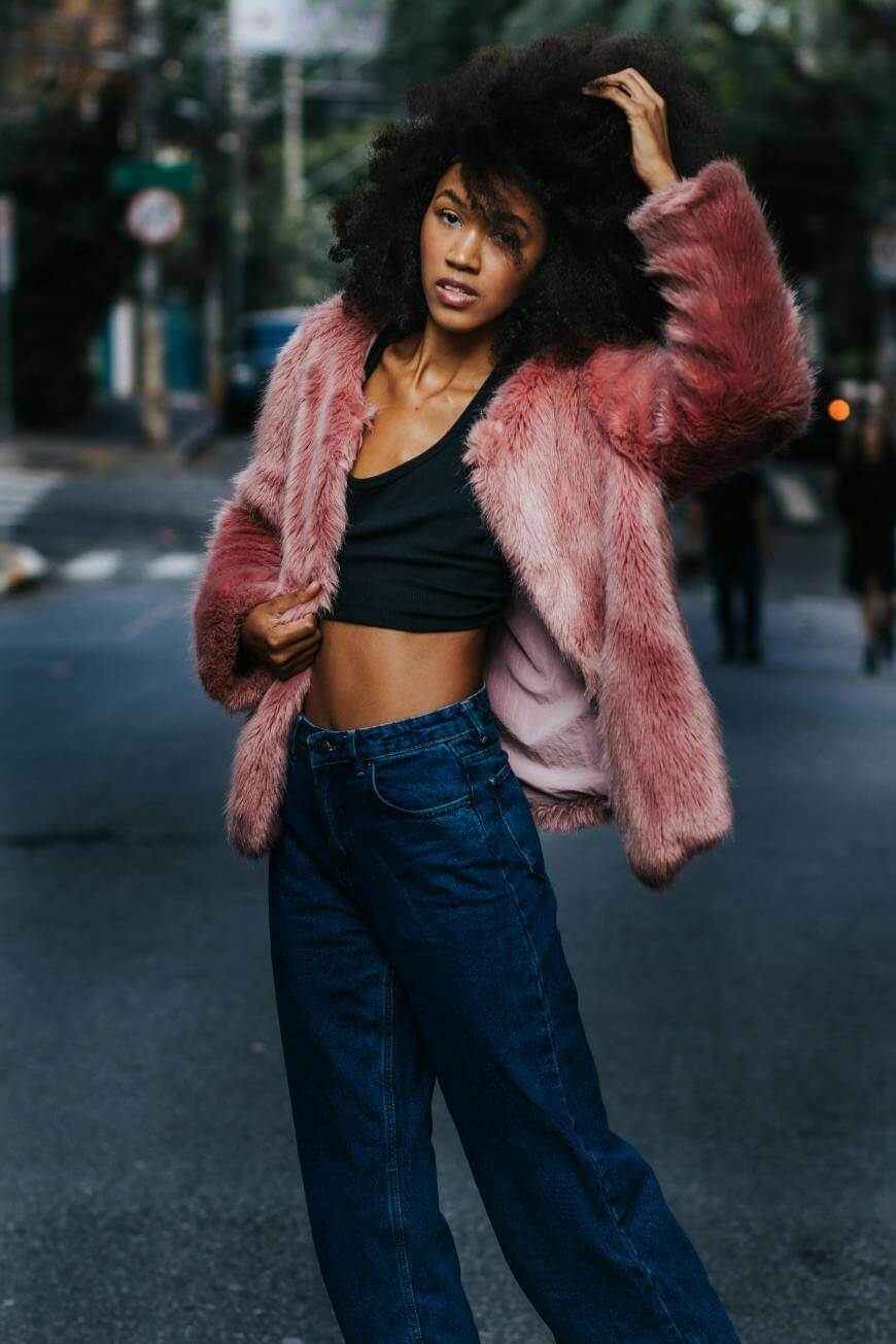A woman stands on a city street wearing a pink faux fur jacket, black crop top, and blue jeans. She touches her curly hair with one hand and looks at the camera, with blurred urban buildings in the background.