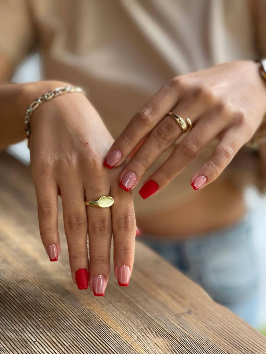 A close-up of two hands with manicured nails; the nails are painted red and light pink in an alternating pattern. The person is wearing gold rings and a gold bracelet, with a blurred background.