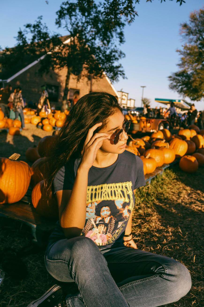 A young person wearing sunglasses and a graphic T-shirt sits among pumpkins at a pumpkin patch on a sunny day, with people and a barn in the background.