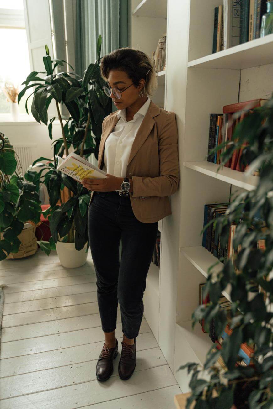 A person wearing glasses, a tan blazer, white shirt, and dark pants stands in a cozy room, leaning against a white bookshelf and reading a book, surrounded by tall green plants.