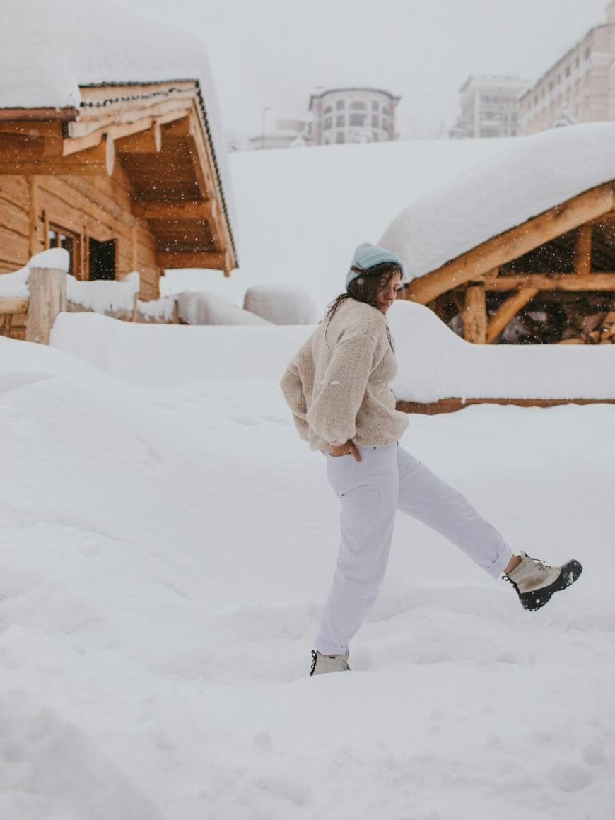 A person dressed in winter clothes walks through deep snow near wooden cabins, with snow-covered roofs in a snowy, mountainous setting.