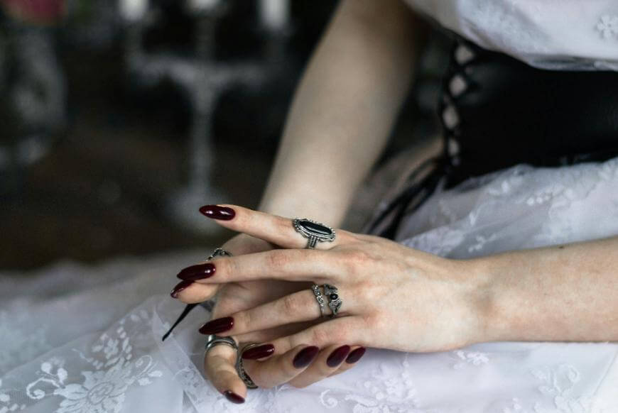 A person wearing a white lace dress rests their hands on their lap, showing dark red polished nails and multiple silver rings with ornate designs. The background is blurred.