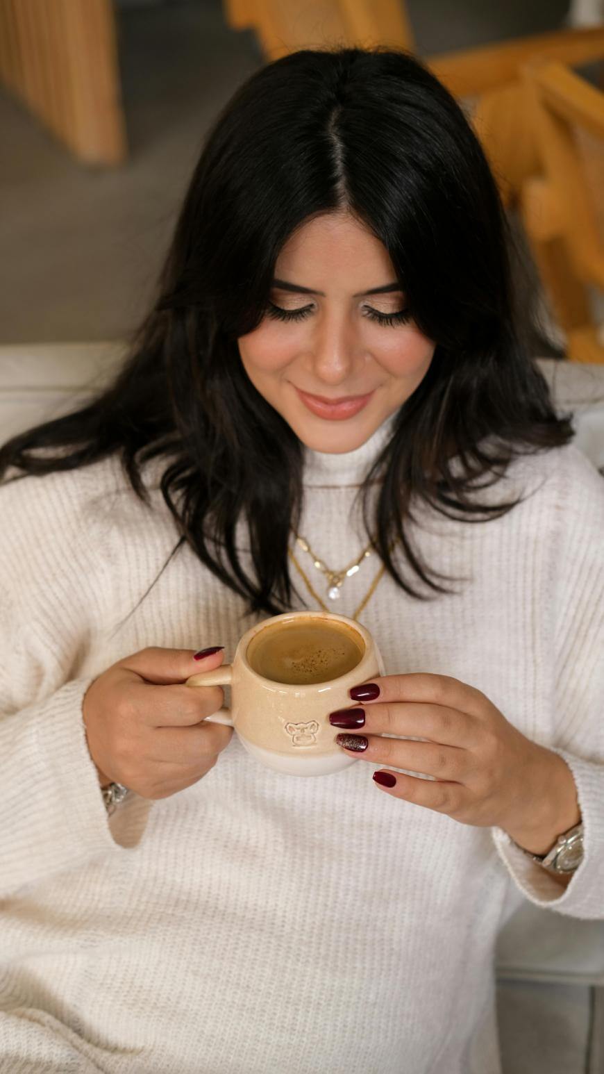 A woman with long dark hair, wearing a white sweater, sits and smiles while holding a beige cup of coffee with both hands. Her nails are painted dark red and she wears a gold necklace and watch.