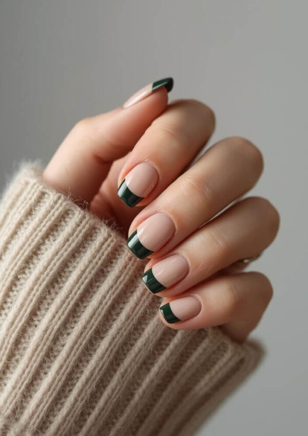 A hand with neatly manicured nails featuring a nude base and dark green French tips, shown against a neutral background. The person is wearing a beige, ribbed-knit sweater.